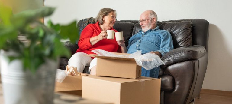 Älteres Paar packt Umzugskartons in einem gemütlichen Wohnzimmer aus. Die Kartons sind mit dem Logo von Umzug MIT Plan versehen, was auf einen organisierten Seniorenumzug hinweist.